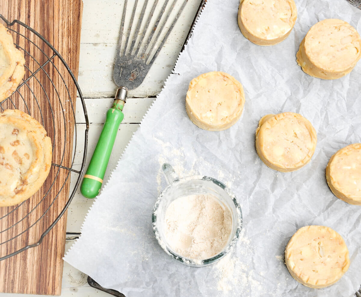 Circles of cookie dough on a baking sheet next to a container of floor, a baking utensil, and baked cookies on a tray