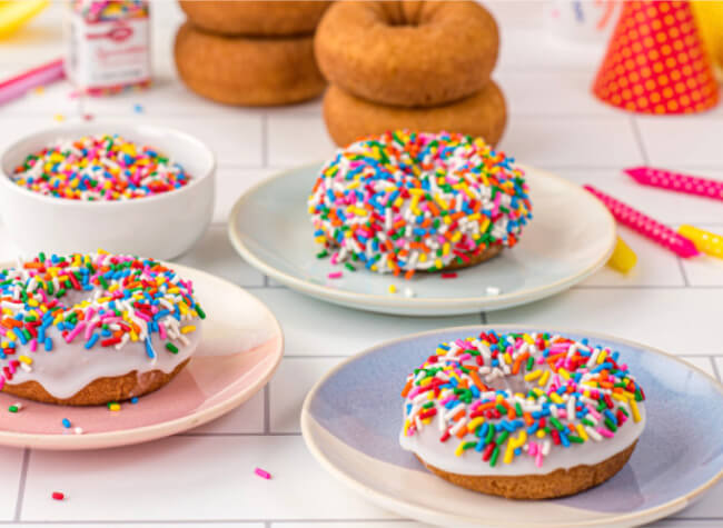 Different donuts with white glaze and multicolored sprinkles on plates with plain donuts and supplies in the background