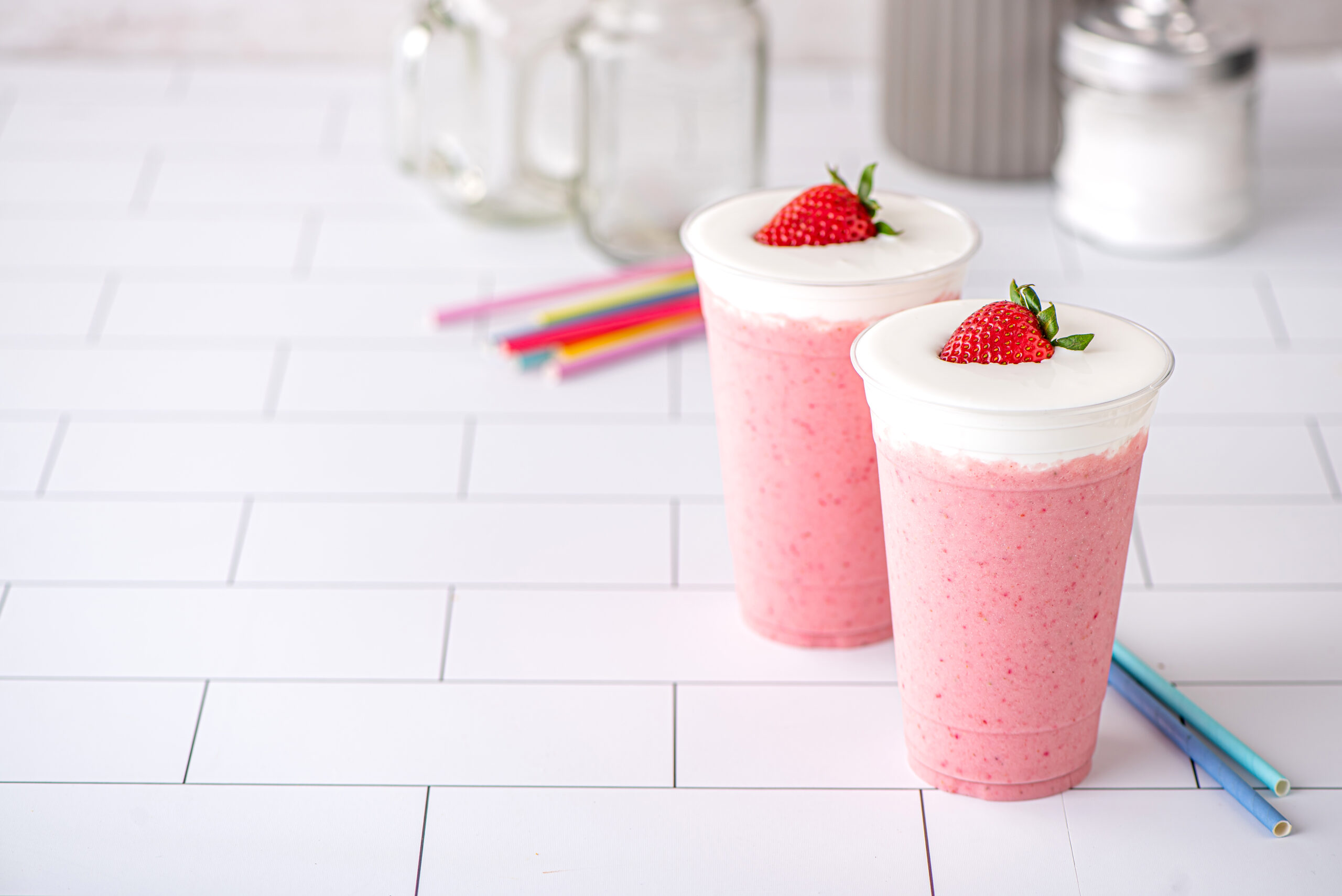Two strawberry milkshakes topped with cream and a strawberry with colorful straws in the background