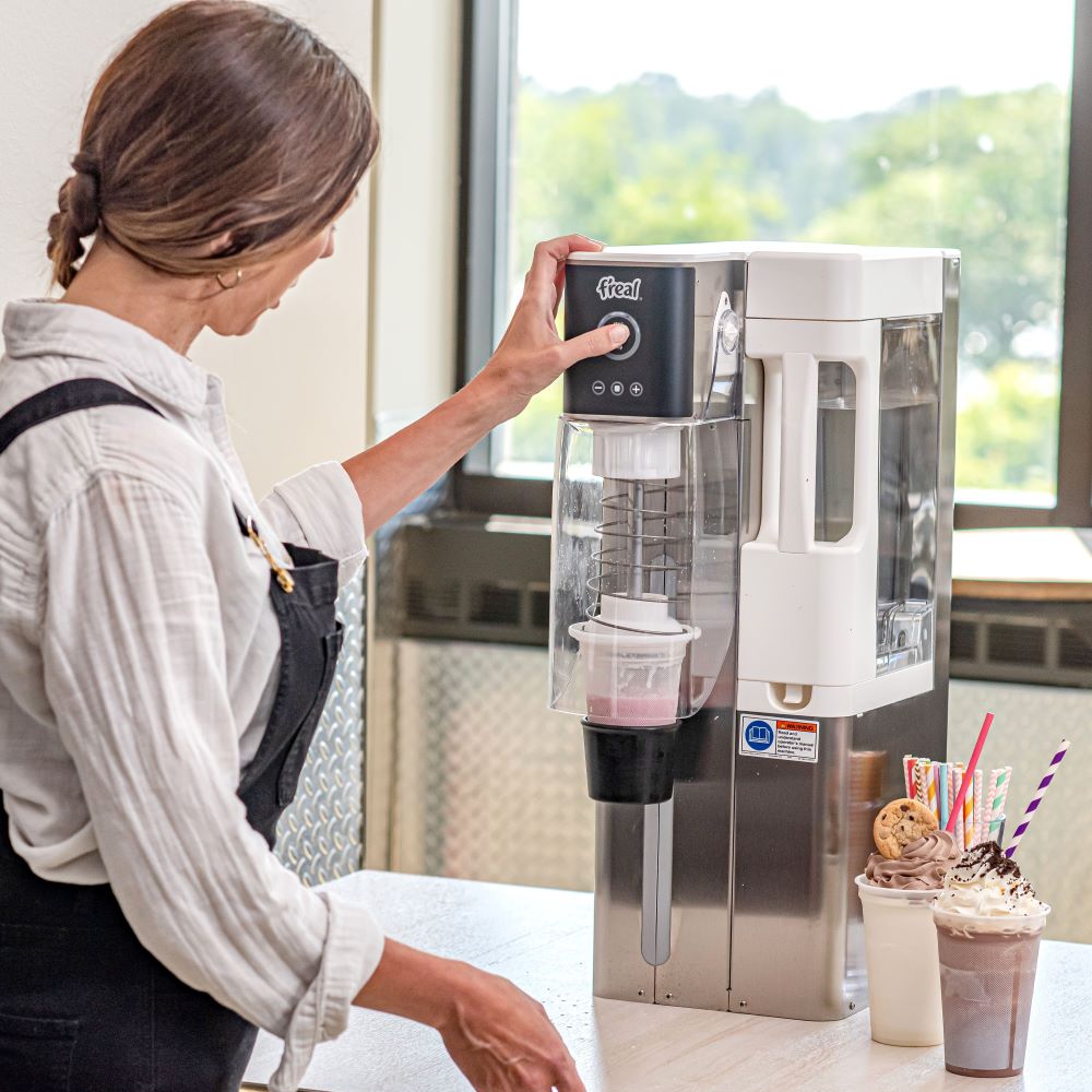A woman wearing a blouse and overalls using a F'real blender machine to make a dessert drink