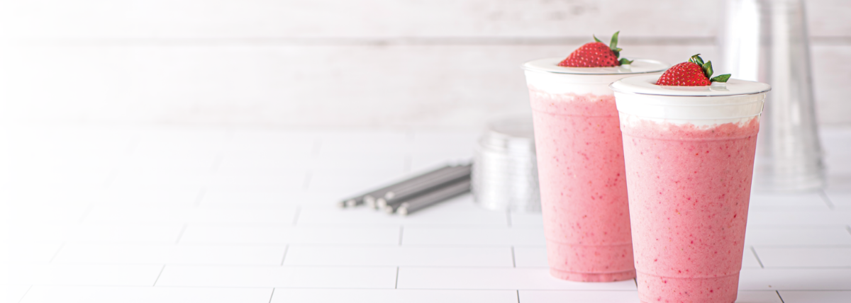Two strawberry drinks topped with cream and a strawberry with silver straws in the background